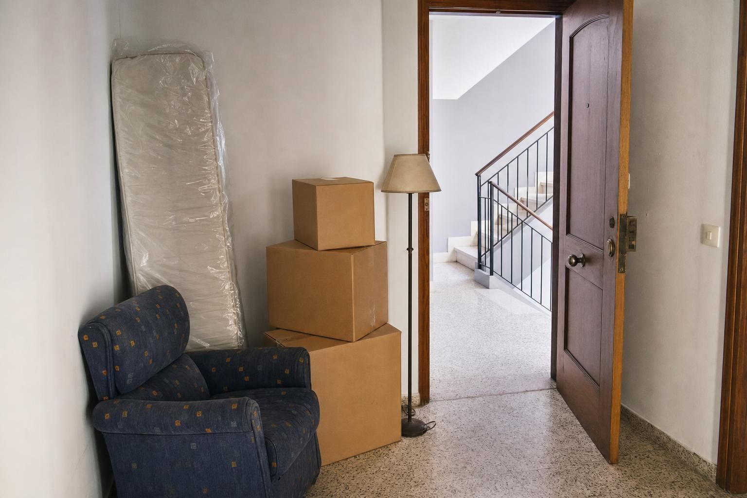 Recibidor de un piso con un sillón, un colchón enfundado y unas cajas junto a la puerta